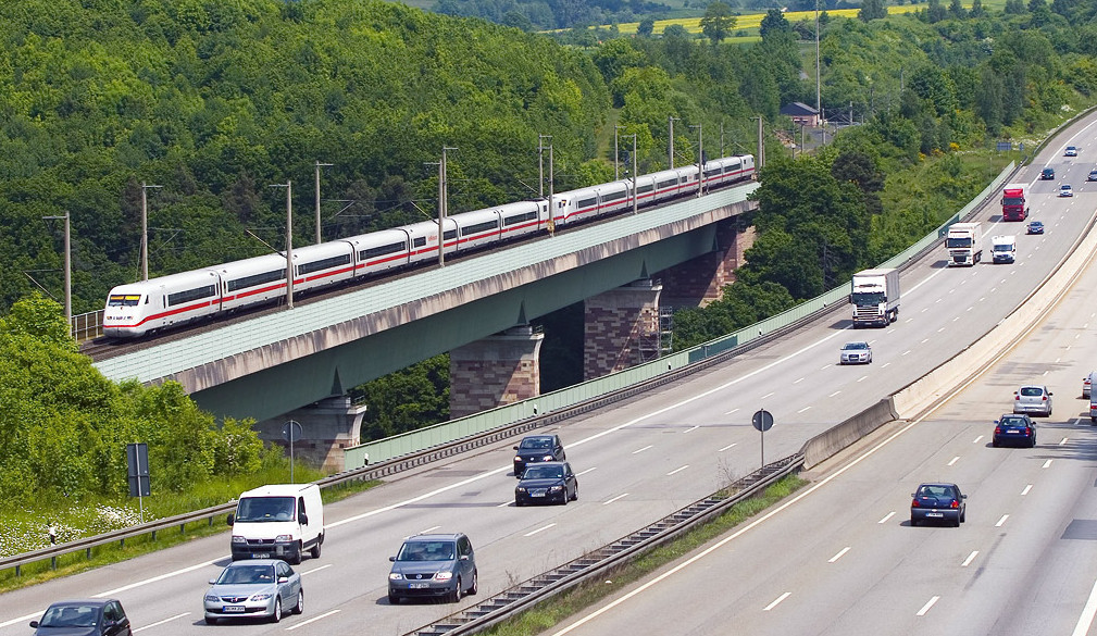 Schnellzug fährt über eine Brücke, im Hintergrund Bäume und Rapsfeld, davor eine Autobahn - Flächenverbrauch